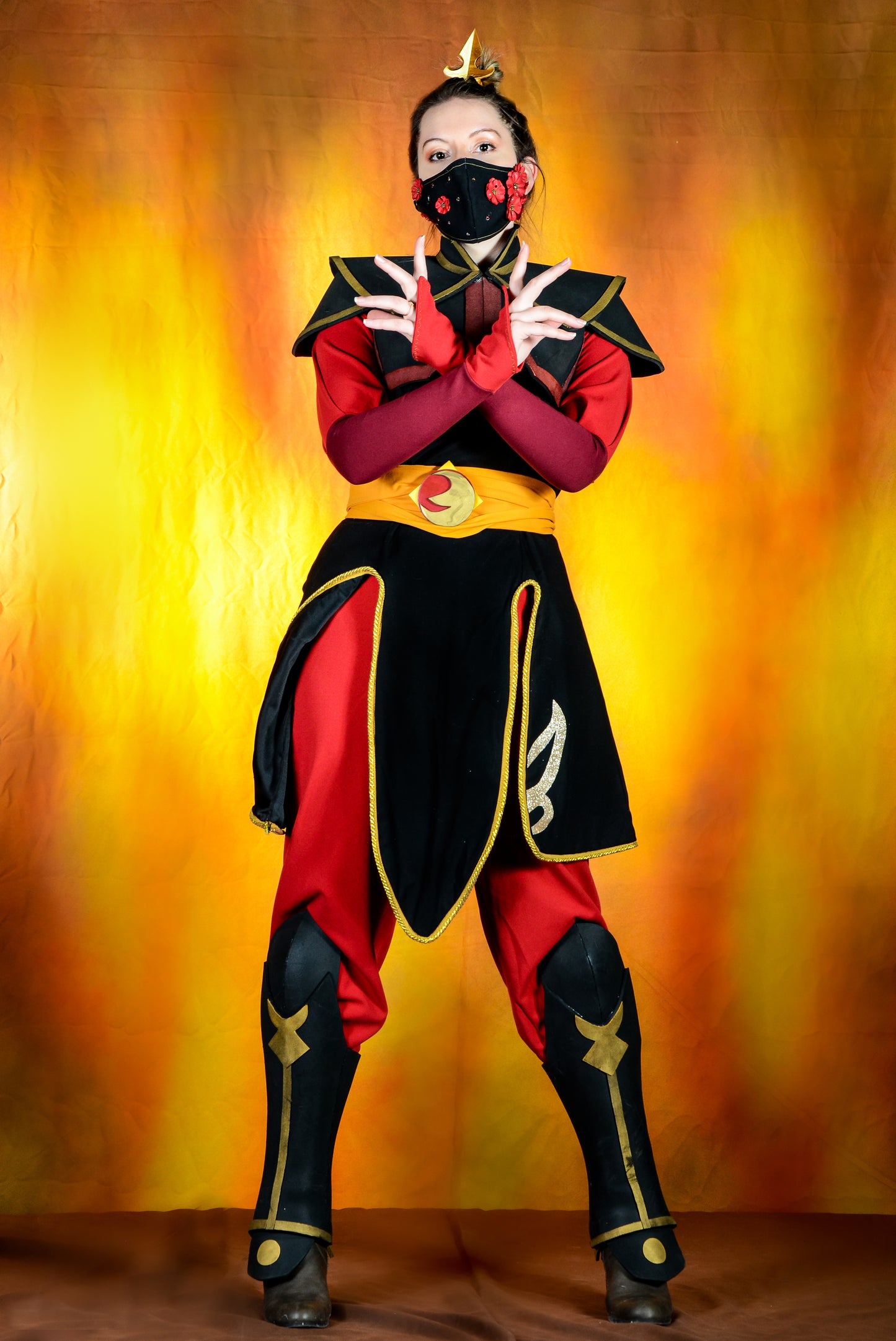 A person wearing a Azula cosplay costume with a crown, shoulder armor, a black tunic with gold trim, a gold belt, and red pants with leg armor, standing against a fiery background.