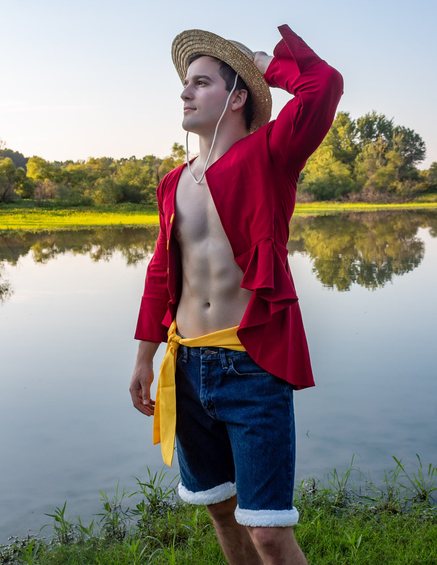 man wearing a luffy inspired cosplay from One Piece. Luffy's New World Outfit. He has a straw hat and red jacket with a yellow belt. He is outside by the water.