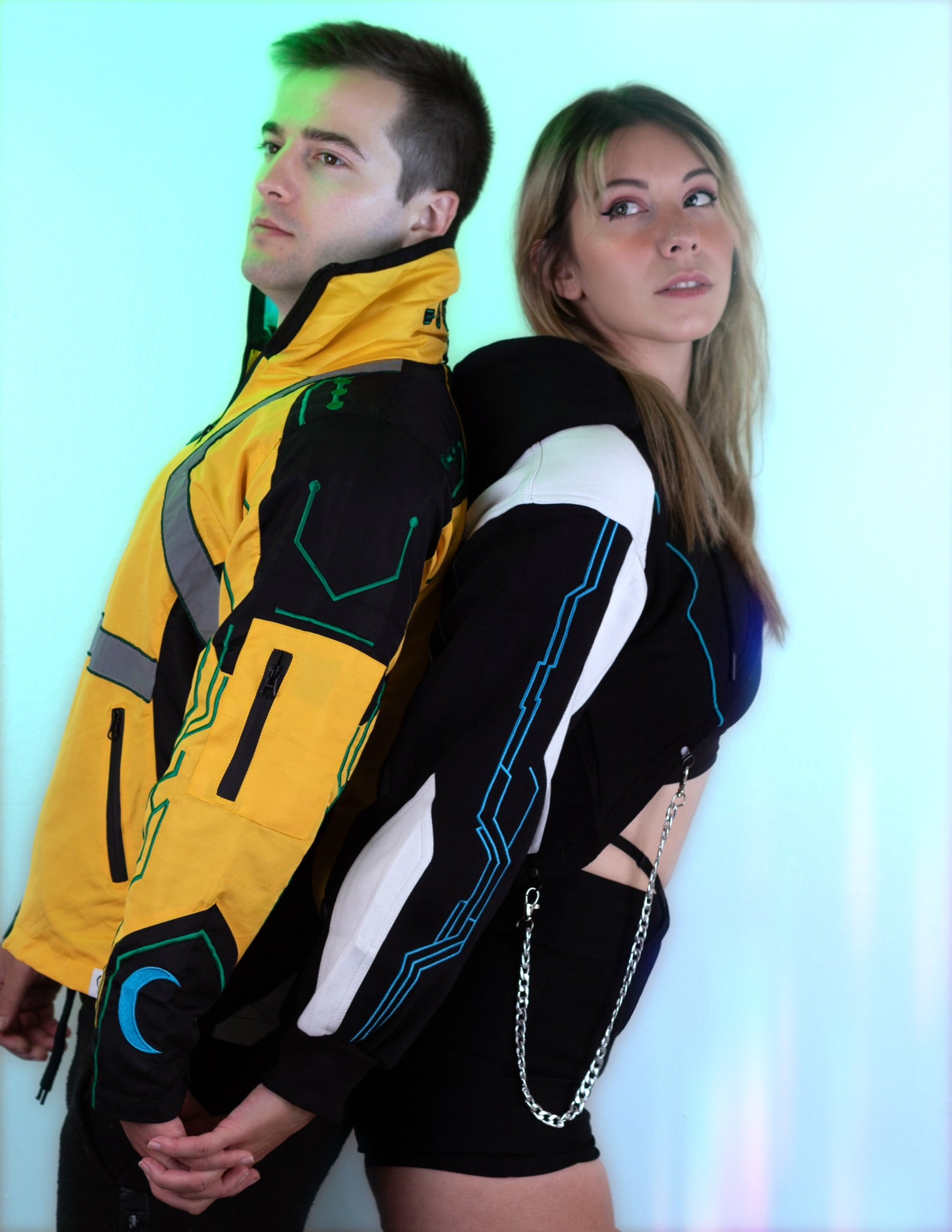 A man and a woman posing back-to-back in futuristic sci-fi themed outfits. The man wears a yellow, black, and gray jacket with green accents, and the woman wears a black crop top with white panels, blue line details, and a silver chain strap.
Inspired by David and Lucy Cyberpunk