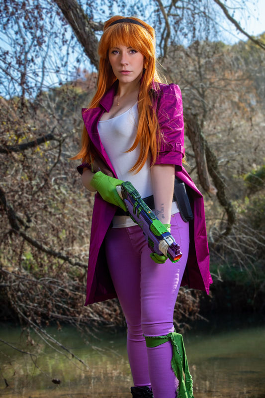 Person in a purple outfit with green gloves and a weapon, standing outdoors near water and trees. Inspired by Daphne from Scooby Doo and the Apocolyptic Comic Book Series.