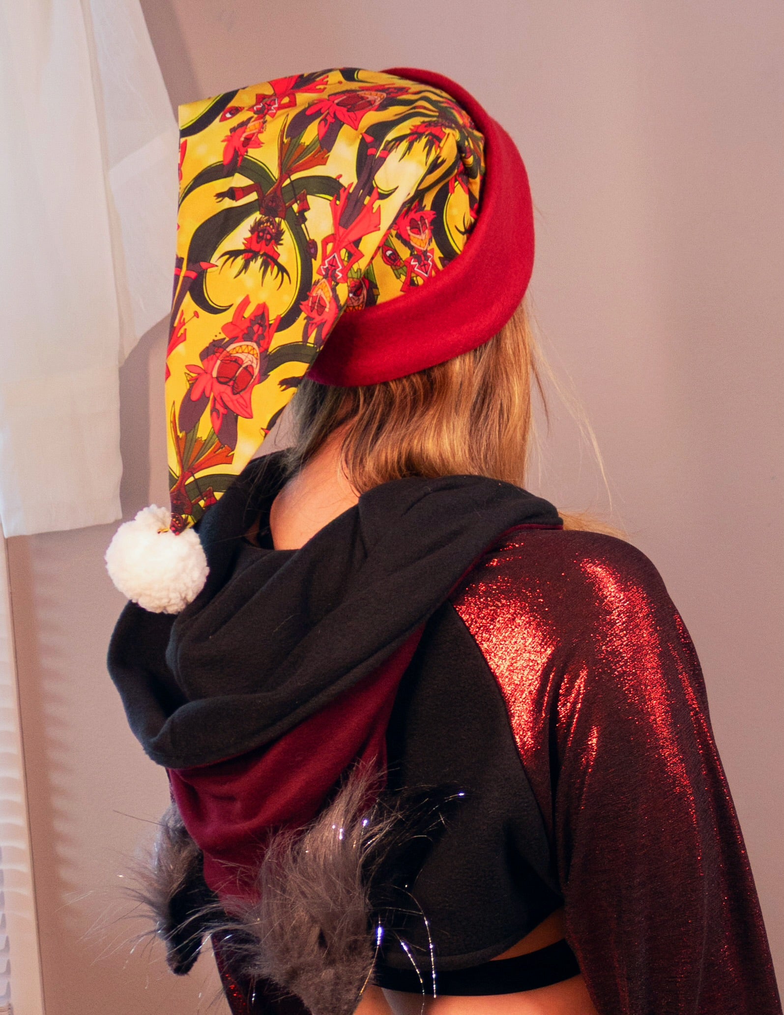 Person wearing a colorful floral-patterned hat and shiny red cosplay shrug indoors. Print is of Alastor from Hazbin hotel.