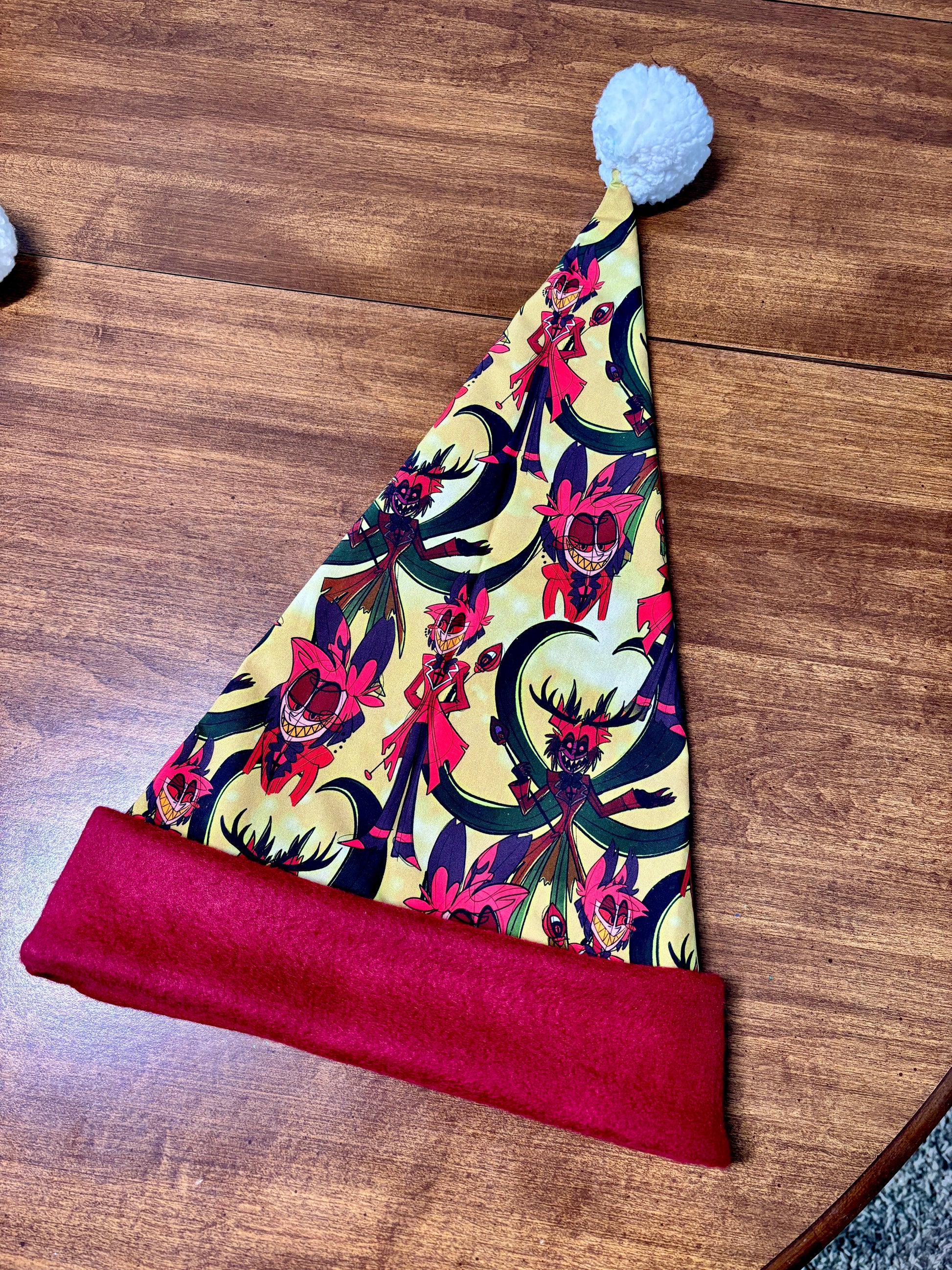 Colorful Santa hat with a red band on a wooden surface. Print is of Alastor from Hazbin hotel.