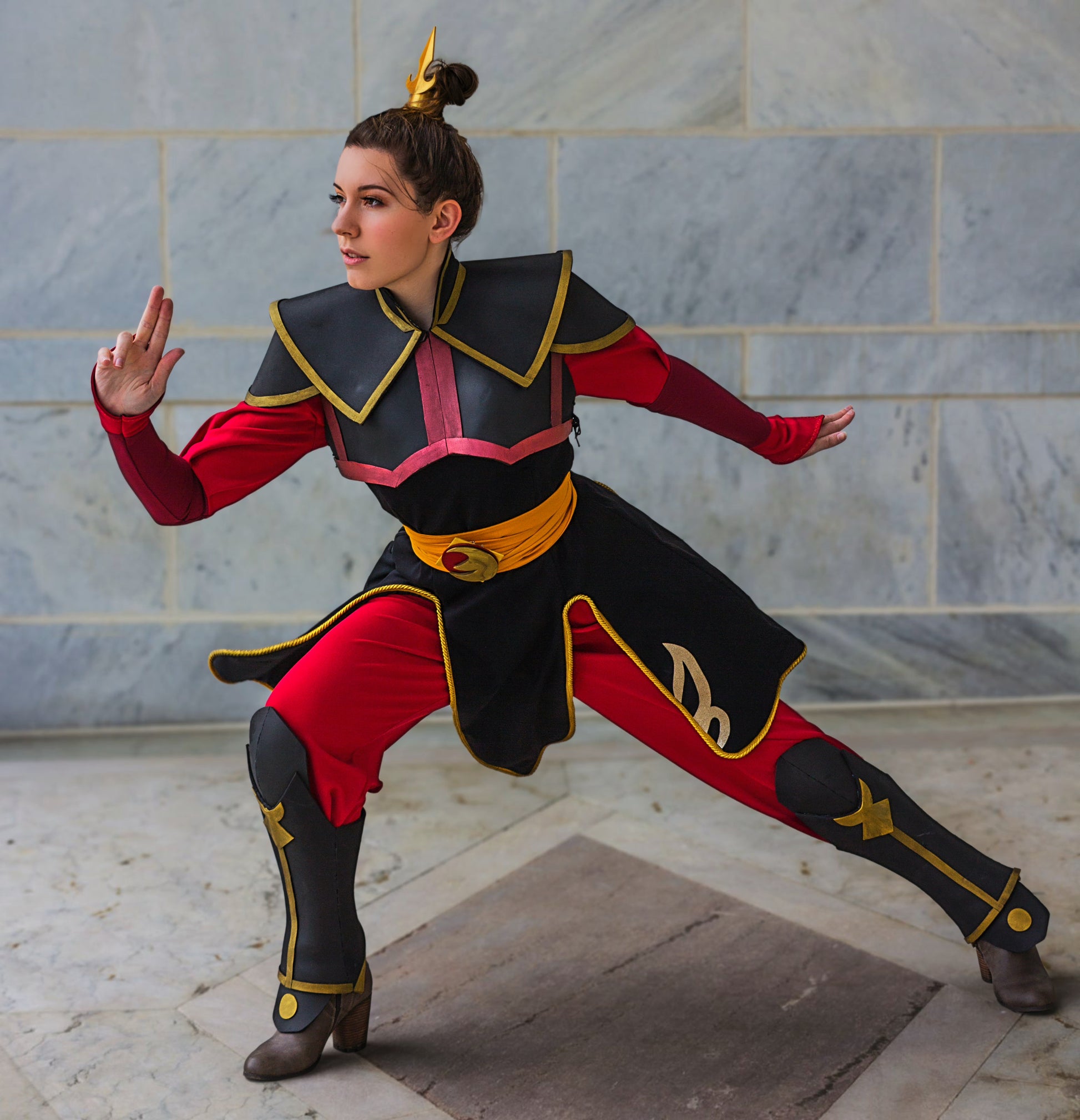 Person in a red and black costume with gold accents posing against a marble wall.