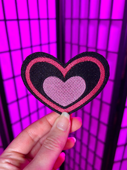 Heart-shaped patch with pink center and black border held against a purple grid background. Heart is inspired by Valentine's Day Fizzarolli from Helluva Boss.