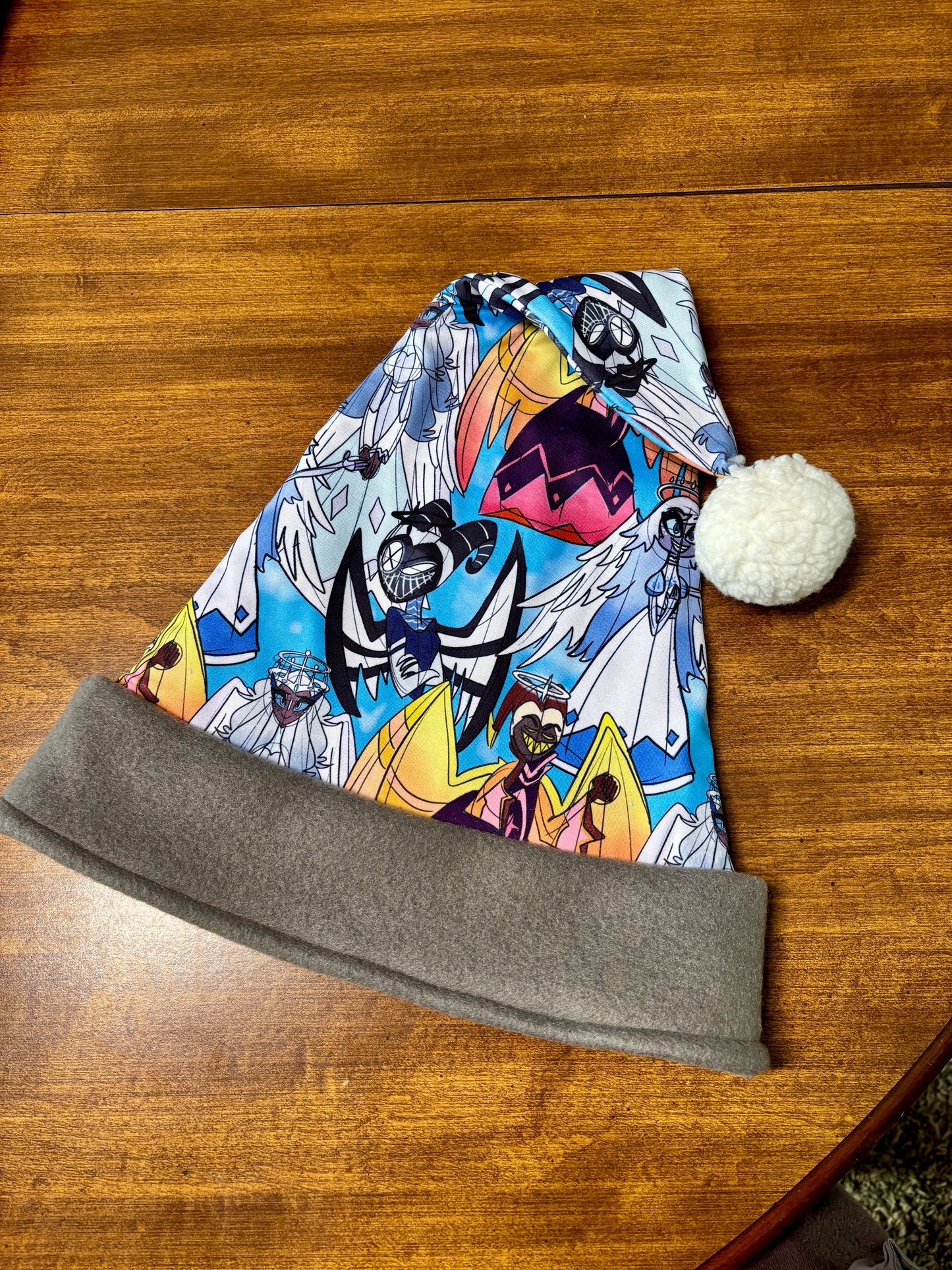 Colorful beanie with character design on a wooden surface. Print  of the Santa hat has Hazbin Hotel characters on it like Adam and Lute.