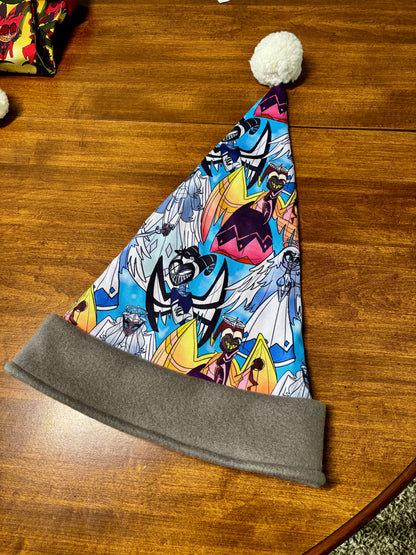 Colorful Santa hat with a gray band on a wooden surface. Print  of the Santa hat has Hazbin Hotel characters on it like Adam and Lute.