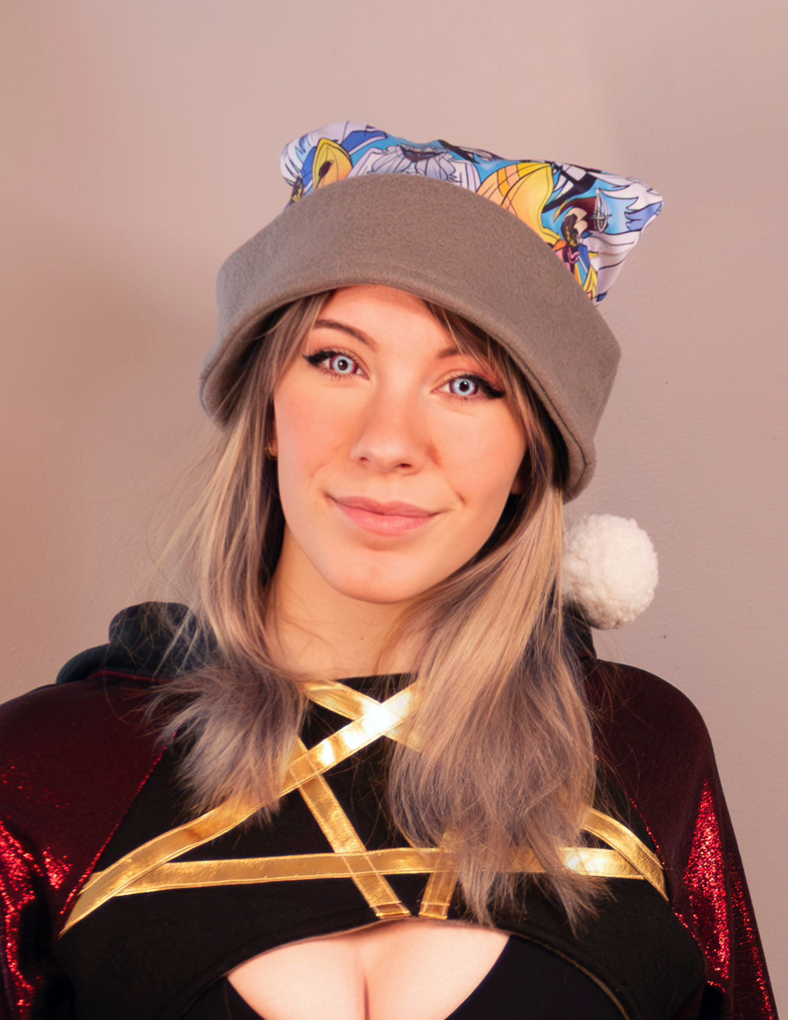 Person wearing a colorful hat with pom-poms on a plain background. Print  of the Santa hat has Hazbin Hotel characters on it like Adam and Lute.