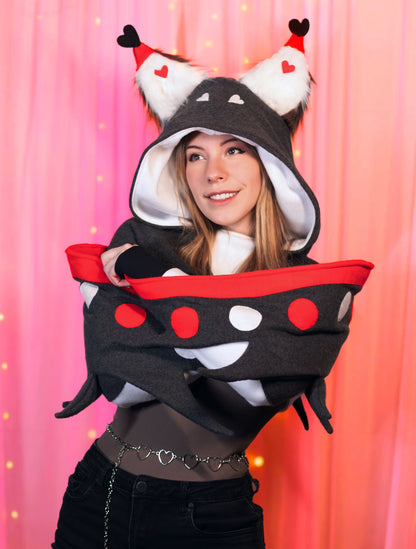 Woman wearing a Husk from Hazbin Hotel inspired shrug against a pink background. She is wrapping the wings around herself to show the softness of the wings.