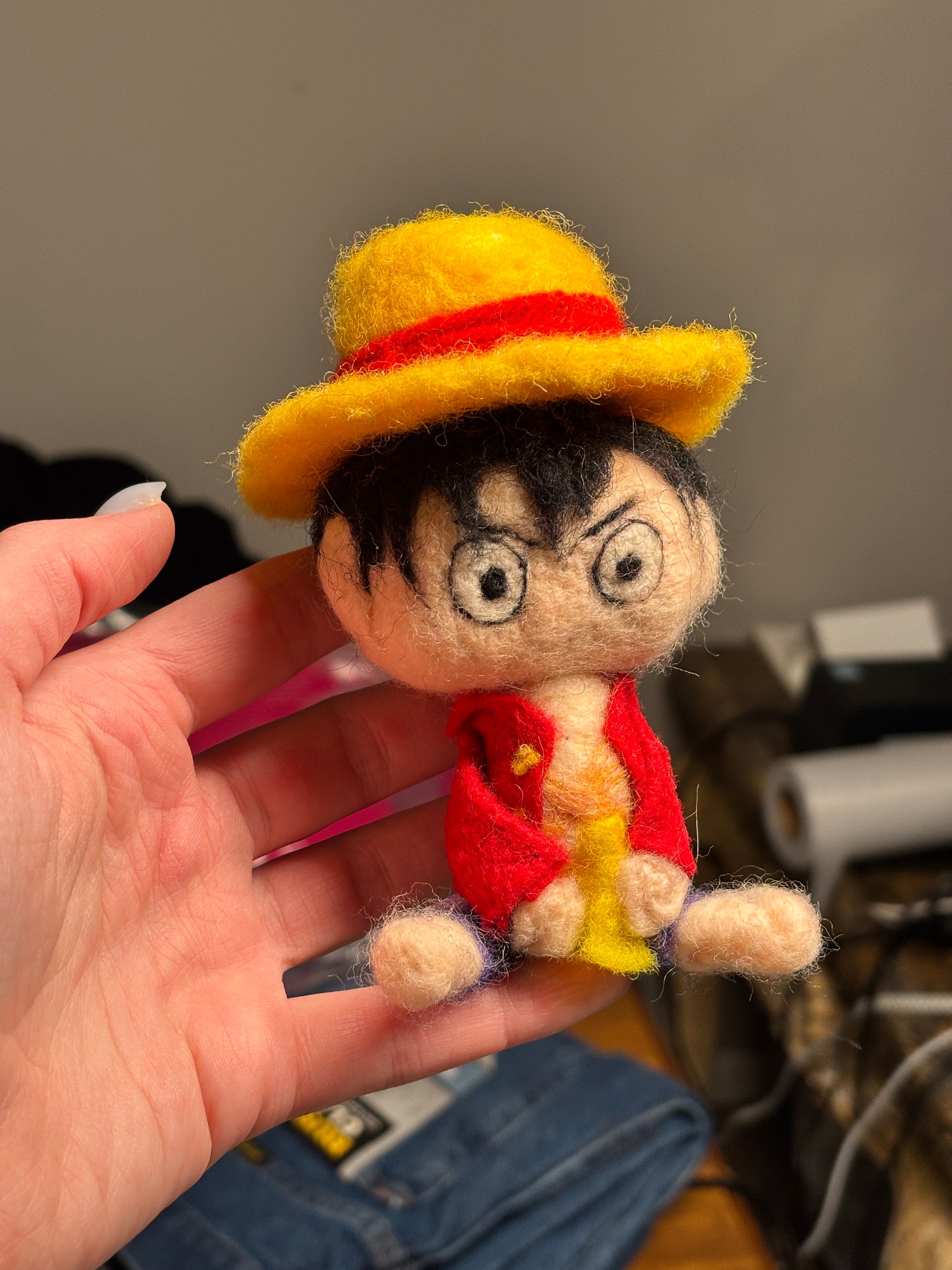 Hand holding a small, colorful felt doll with a yellow hat and red accents. Inspired by Luffy from One Piece. 
