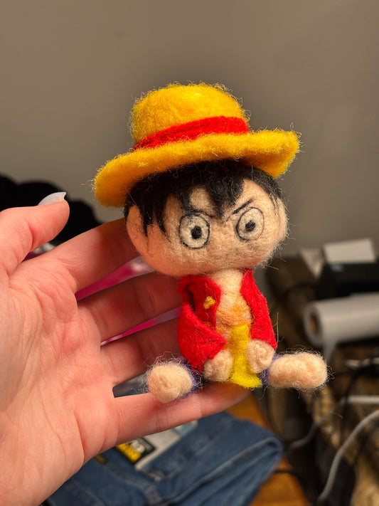 Hand holding a small, colorful felt doll with a yellow hat and red accents. Inspired by Luffy from One Piece. 