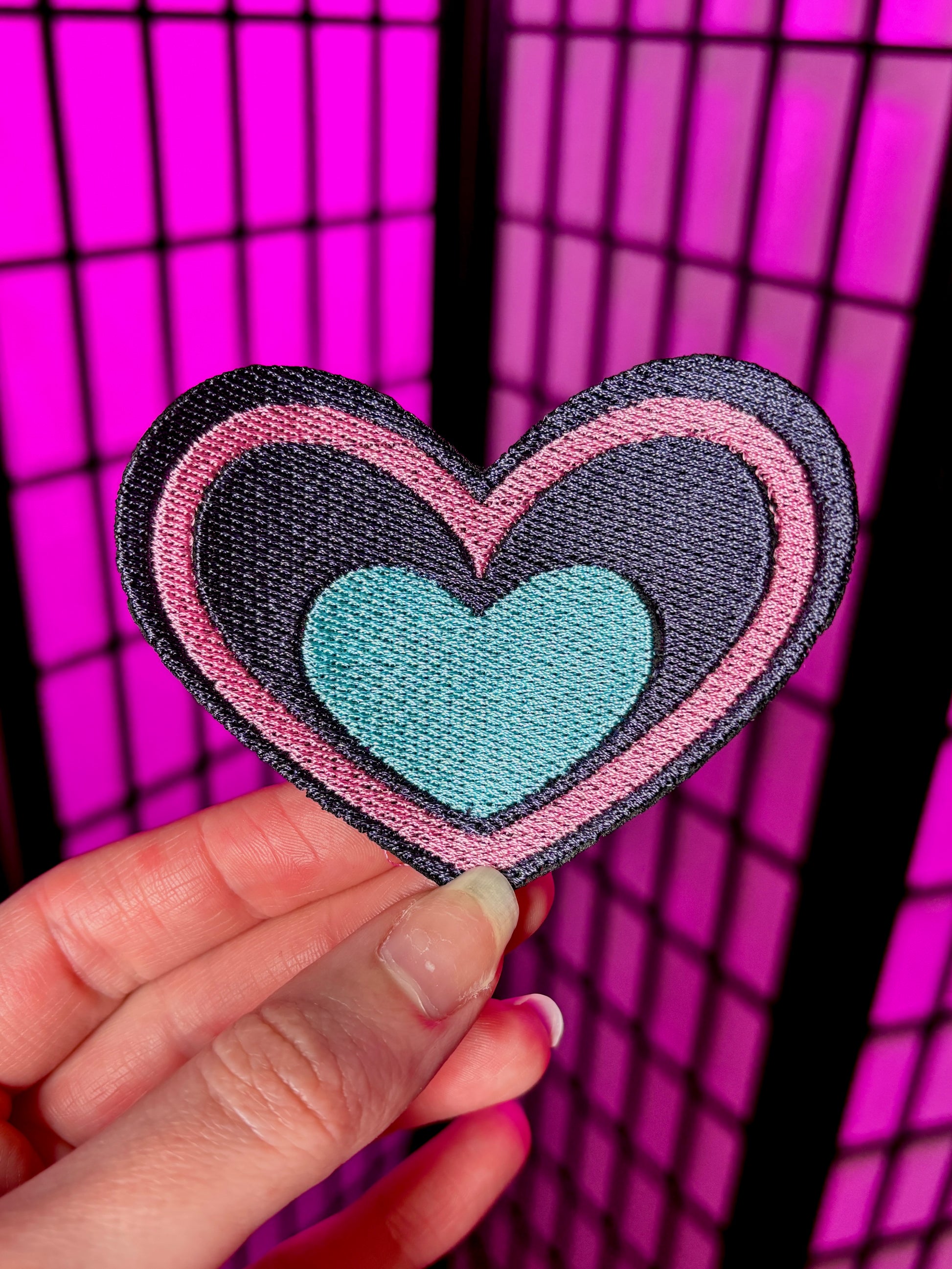 Heart-shaped patch with a turquoise center and pink and black outline held against a pink grid background. Inspired by Pastel Fizzarolli from Helluva Boss.
