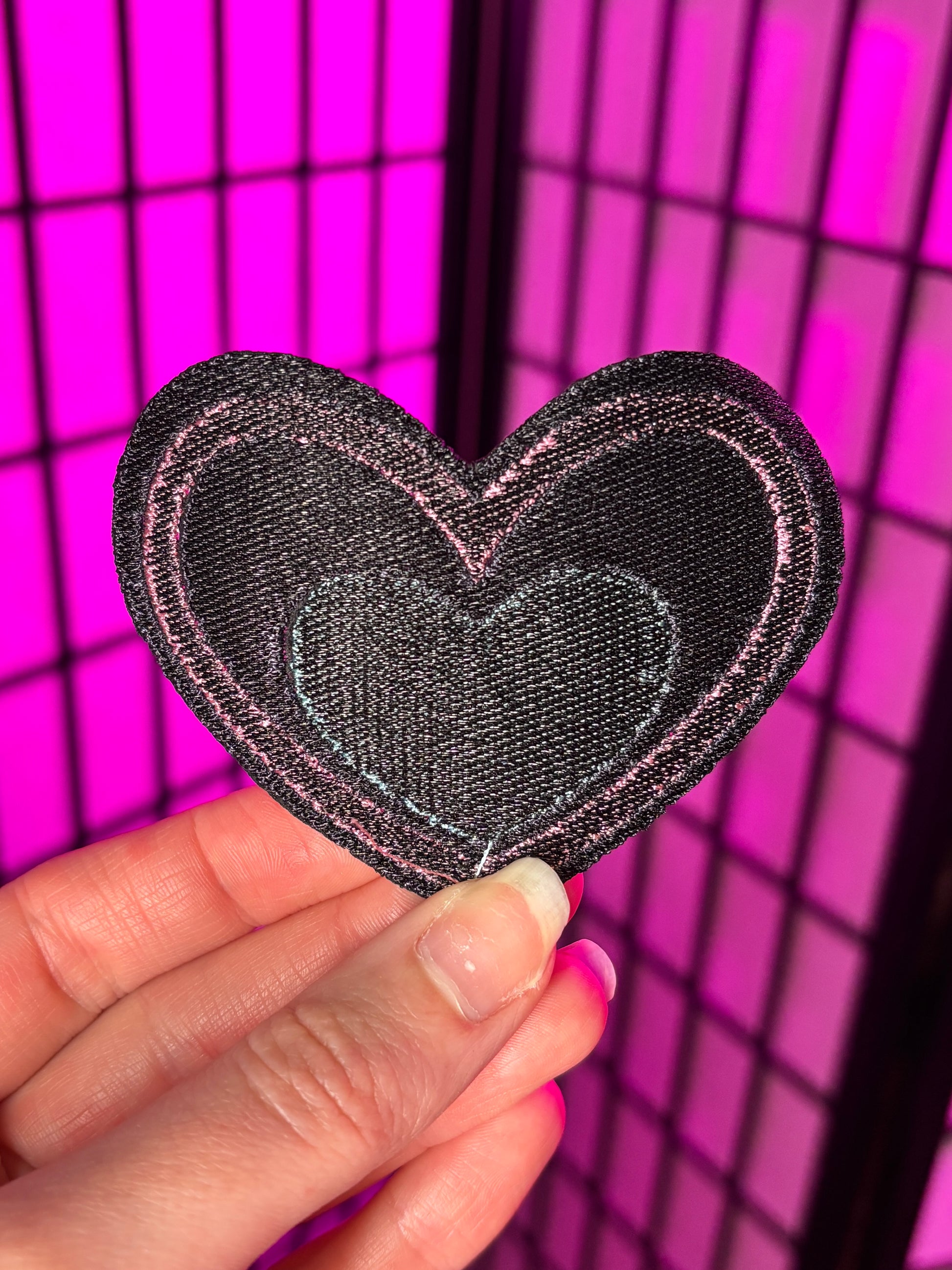 Heart-shaped patch held by a hand against a pink grid background. Showing the back of the patch which is dark thread. It does not have addhesive. Inspired by Pastel Fizzarolli from Helluva Boss.