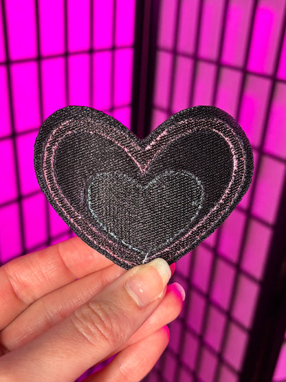 Heart-shaped patch held by a hand against a pink grid background. Showing the back of the patch which is dark thread. It does not have addhesive. Inspired by Pastel Fizzarolli from Helluva Boss.