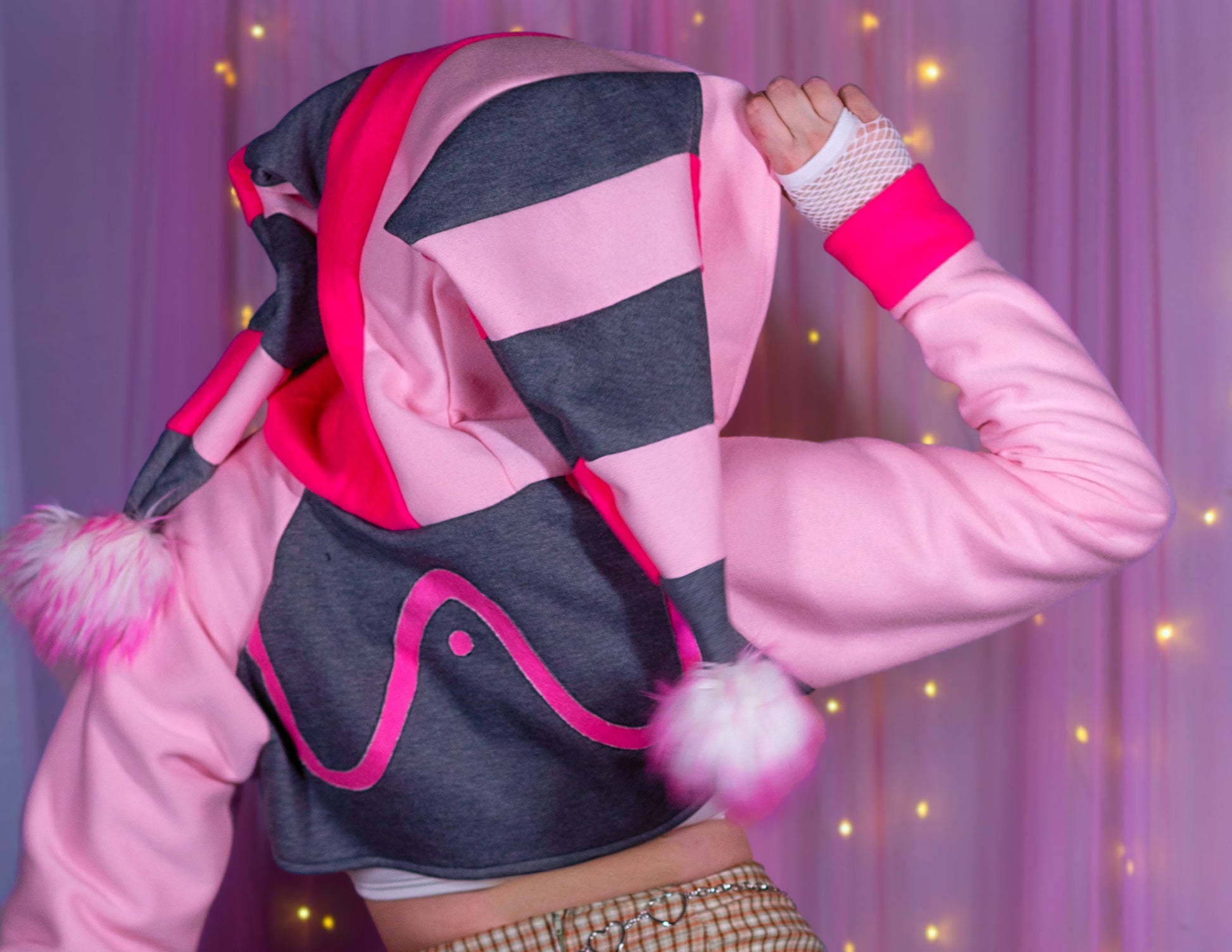 Woman wearing a Valentine's Day shrug inspired by Fizzarolli from Helluva Boss. There is a pink background with twinkle lights. She is wearing a kawaii matching outfit. Showing the back of the shrug with an emphasis on the pink tipped white poms.