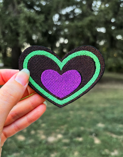 Heart-shaped patch with green border and purple center held by a hand outdoors. Inspired by Beetlejuice and Fizzarolli from Helluva Boss