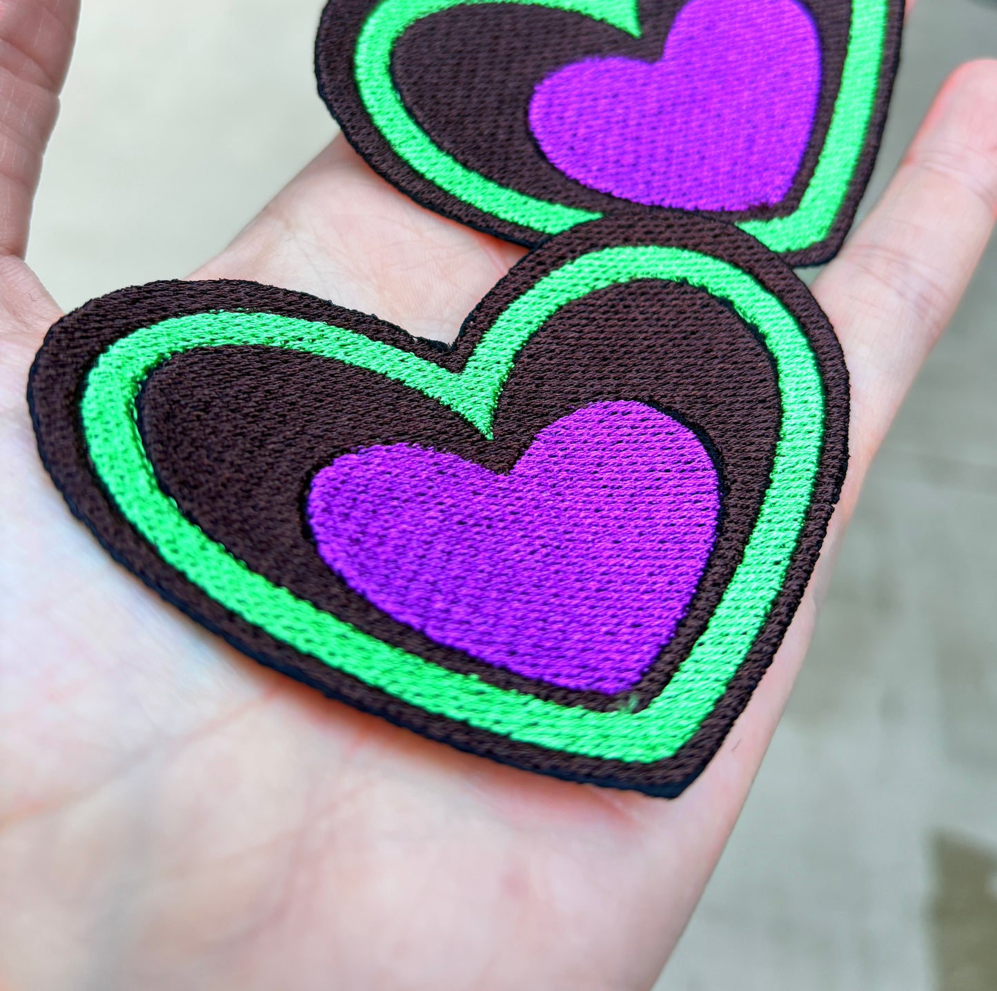 Heart-shaped patch with green border and purple center held by a hand outdoors. Inspired by Beetlejuice and Fizzarolli from Helluva Boss