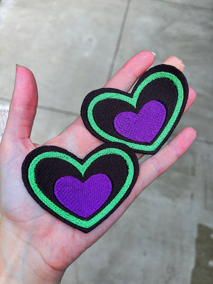 two Heart-shaped patch with green border and purple center held by a hand outdoors. Inspired by Beetlejuice and Fizzarolli from Helluva Boss
