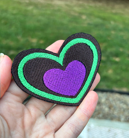 Heart-shaped patch with green border and purple center held by a hand outdoors. Inspired by Beetlejuice and Fizzarolli from Helluva Boss