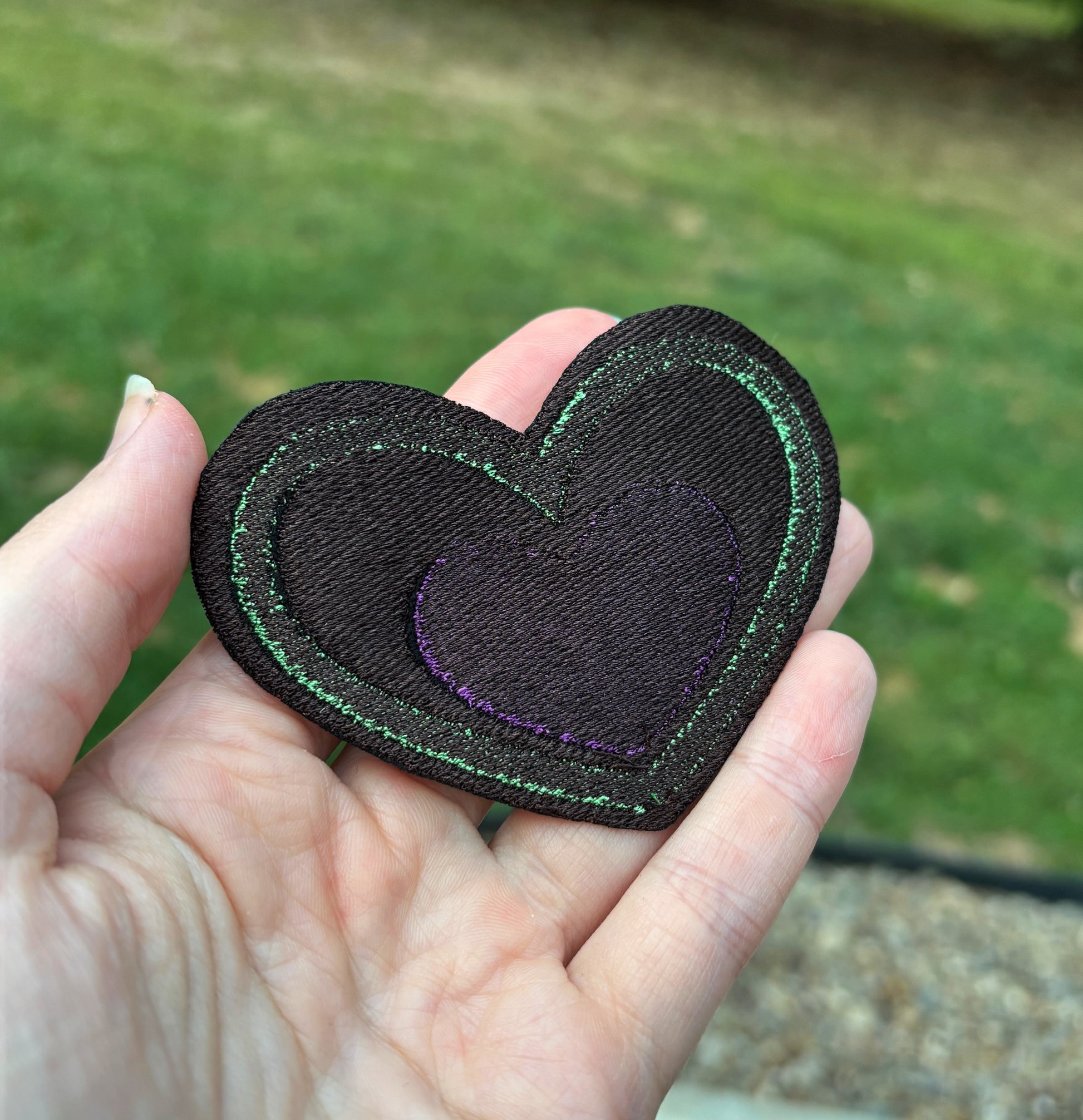 back of a Heart-shaped patch with green border and purple center held by a hand outdoors. Inspired by Beetlejuice and Fizzarolli from Helluva Boss