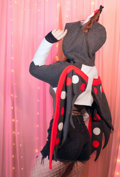 Woman wearing a Husk from Hazbin Hotel inspired shrug against a pink background. Focus is on the back of the shrug shouwing the wings.