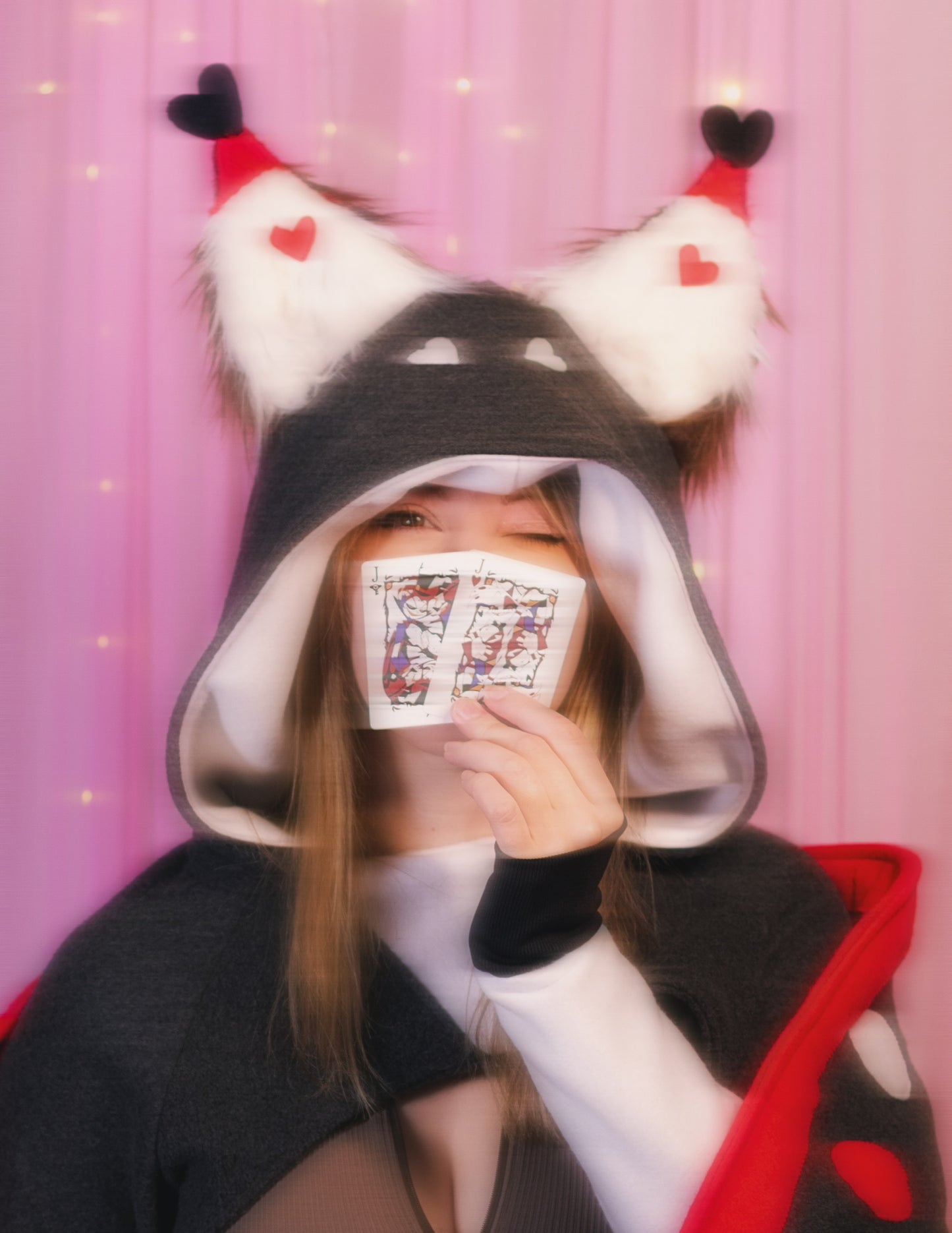 Woman wearing a Husk from Hazbin Hotel inspired shrug against a pink background. Focus is a close up photo of the face and she is holding two classic playing cards displaying prints of Husk and Angel dust from Hazbin Hotel.