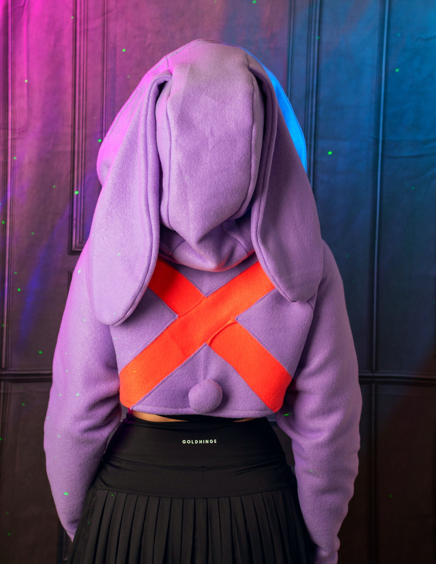 Person wearing a purple hoodie with bunny ears and an orange 'X' design, standing against a dark background. Inspired by Jax from the Amazing digital circus
