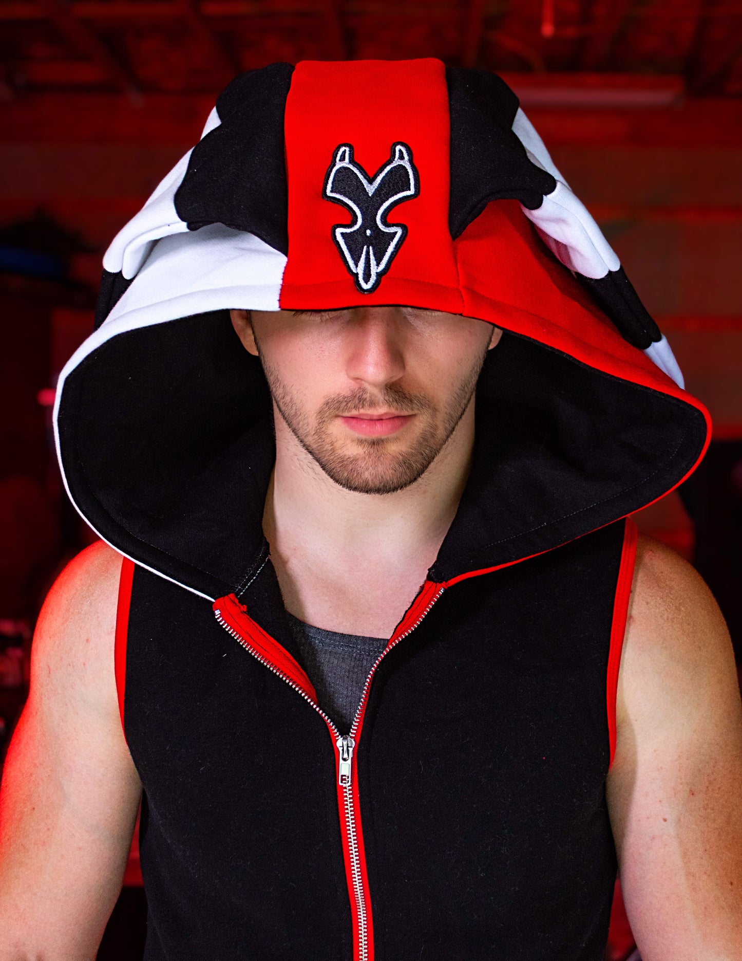 Person wearing a black, red, and white hoodie with a logo on a blurred background. Blitz cosplay inspired by Helluva Boss. sleevelss hoodie