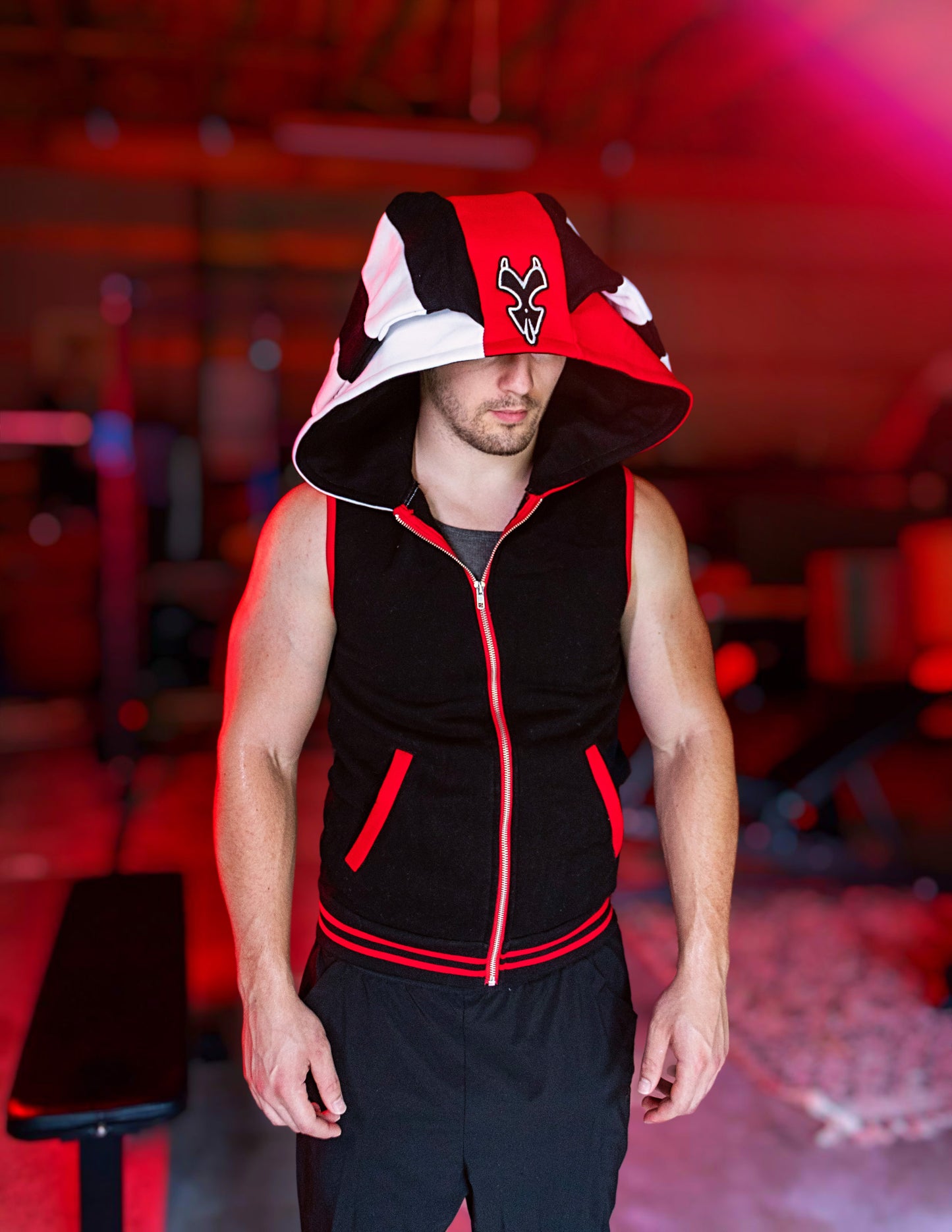 Person wearing a black, red, and white hoodie with a logo on a blurred background. Blitz cosplay inspired by Helluva Boss. sleevelss hoodie