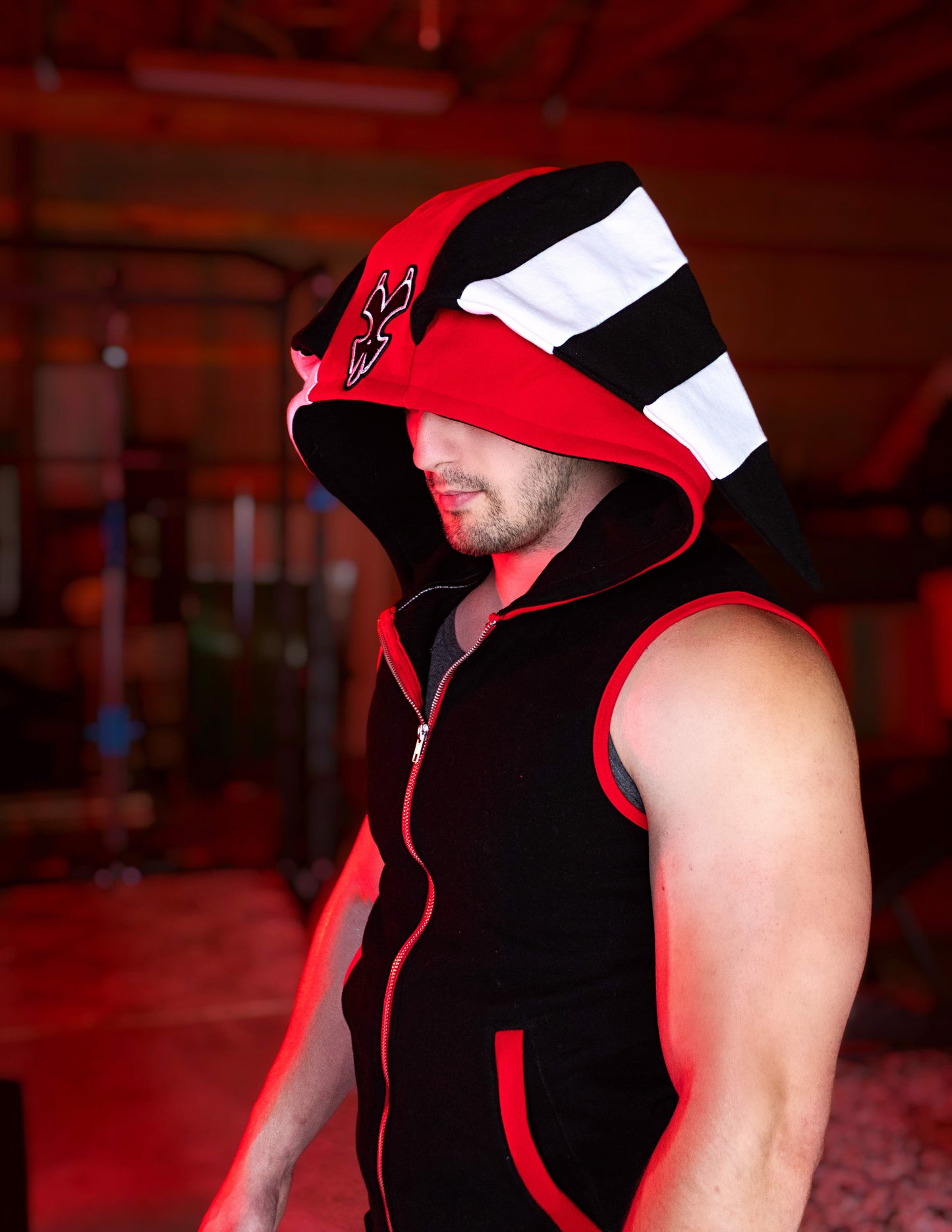 Person wearing a black, red, and white hoodie with a logo on a blurred background. Blitz cosplay inspired by Helluva Boss. sleevelss hoodie