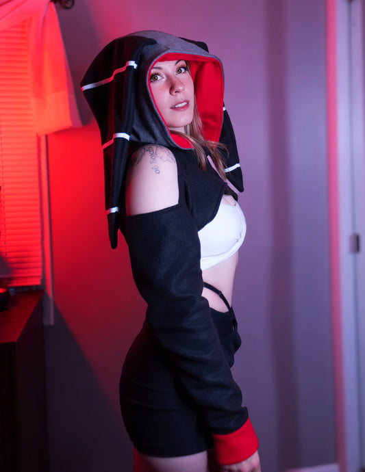 Person wearing a black and red outfit with a hood, standing against a red and gray background. Millie cosplay cropped hoodie shrug inspired by Millie from Helluva Boss