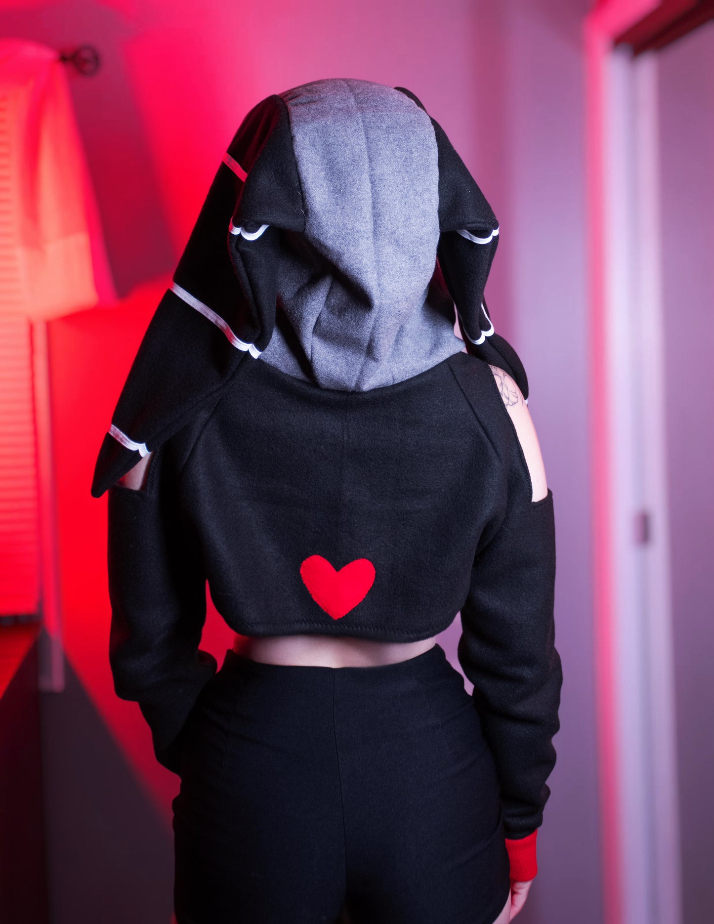 Person wearing a black and red outfit with a hood, standing against a red and gray background. Millie cosplay cropped hoodie shrug inspired by Millie from Helluva Boss
