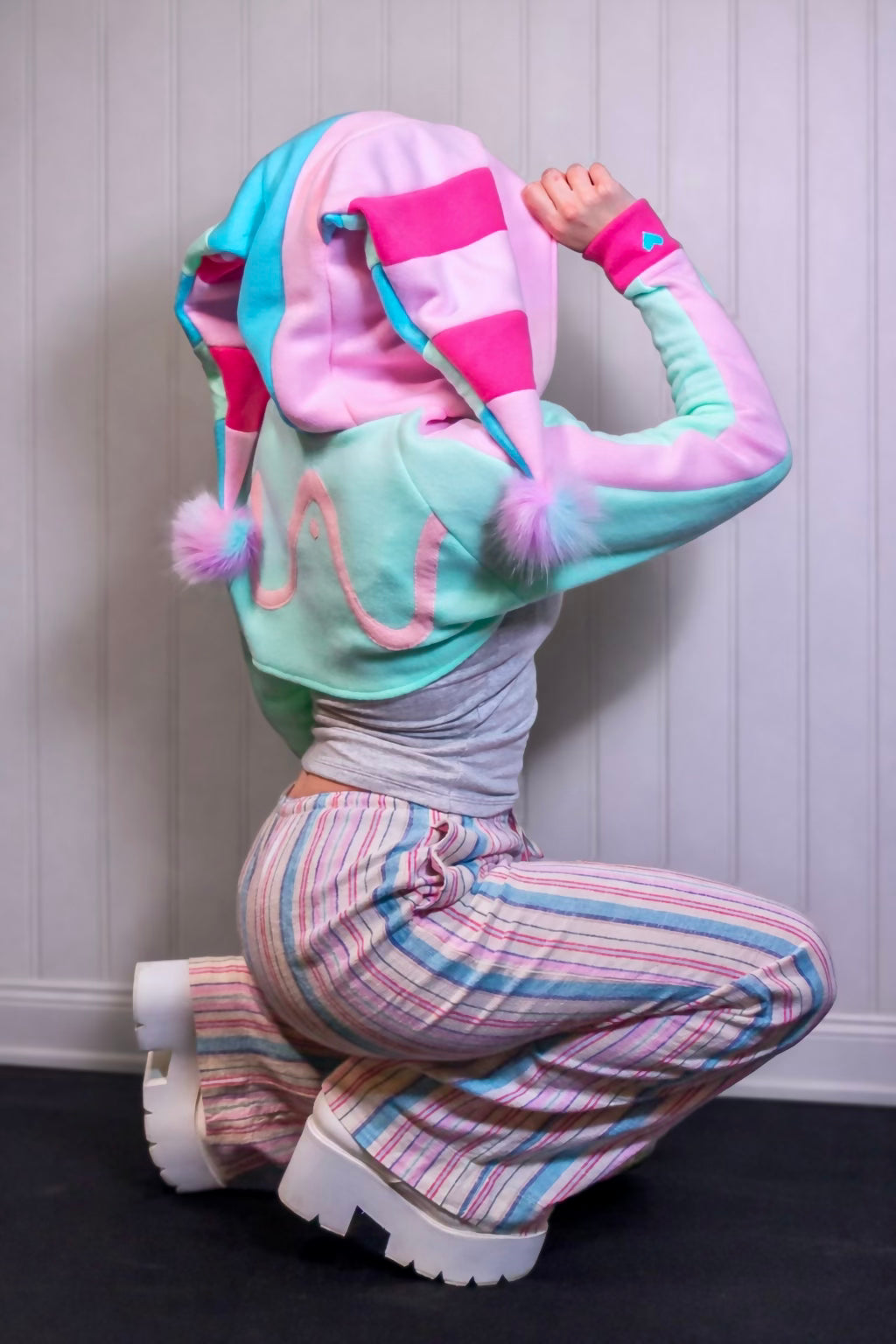 Person wearing a colorful hoodie and striped pants crouching against a white paneled wall. Hoodie is inspired by Fizzarolli from helluva boss in pastel colors