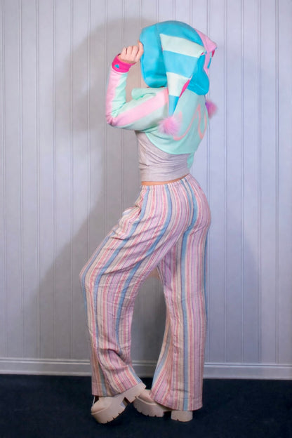 Person wearing a colorful outfit with a pastel striped hoodie and pants against a white paneled wall. cropped hoodie shrug is inspired by Fizzarolli from Helluva Boss in pastel colros.