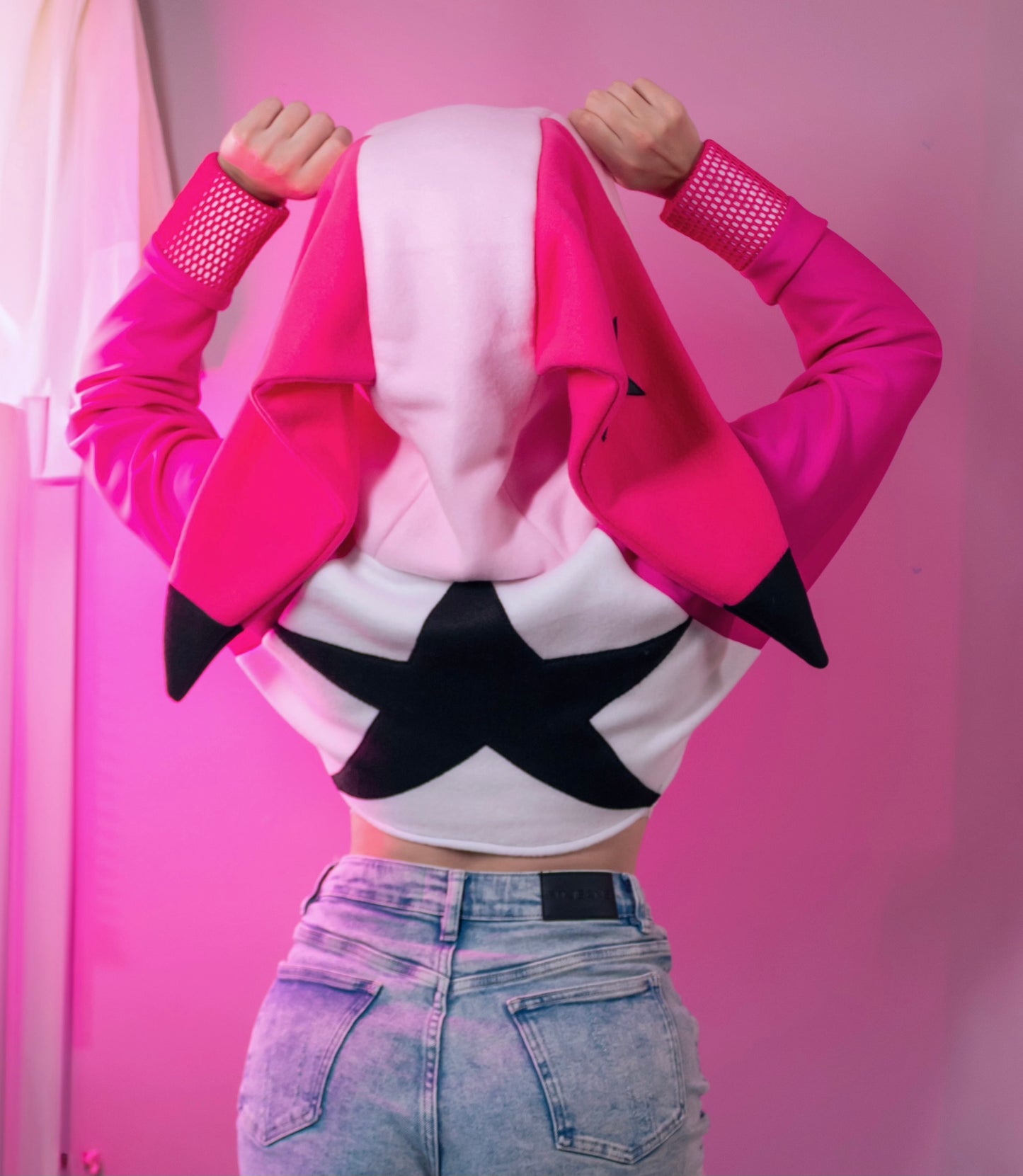 Person wearing a pink cosplay crop hoodie shrug with a hood, white crop top, and denim shorts against a pink background. Inspired by Verosika from Helluva Boss.