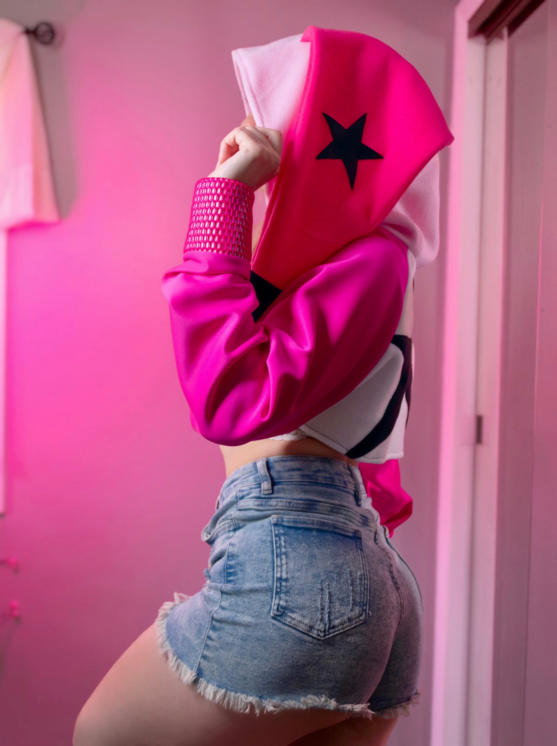 Person wearing a pink cosplay crop hoodie shrug with a hood, white crop top, and denim shorts against a pink background. Inspired by Verosika from Helluva Boss.