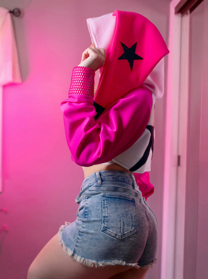 Person wearing a pink cosplay crop hoodie shrug with a hood, white crop top, and denim shorts against a pink background. Inspired by Verosika from Helluva Boss.