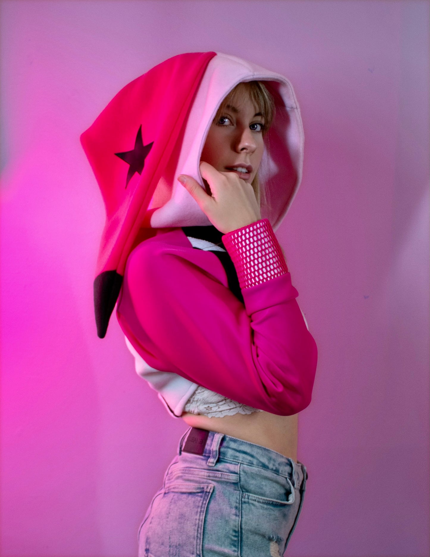 Person wearing a pink cosplay crop hoodie shrug with a hood, white crop top, and denim shorts against a pink background. Inspired by Verosika from Helluva Boss.