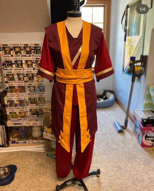 A Zuko cosplay costume set featuring a lined red robe with gold trim, an elastic pants, and a gold belt, displayed on a mannequin in a home setting with various collectibles around.